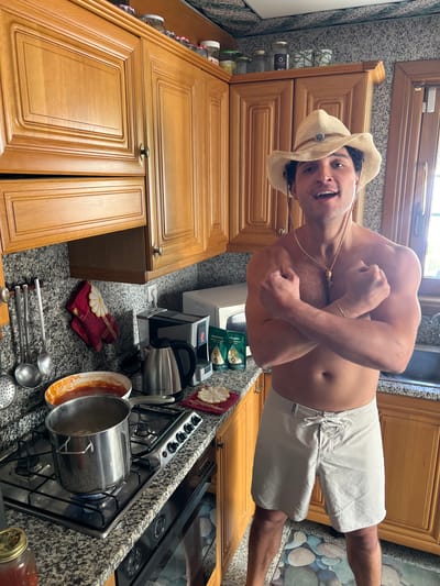 A shirtless man wearing a cowboy hat and white shorts is posing in a kitchen. The kitchen features wooden cabinets, a granite countertop with a stove, and various kitchen items like a kettle, pots, and cooking utensils. The image's location in Èze, France, is not visually confirmed, as there are no elements clearly indicating this specific geographical area.