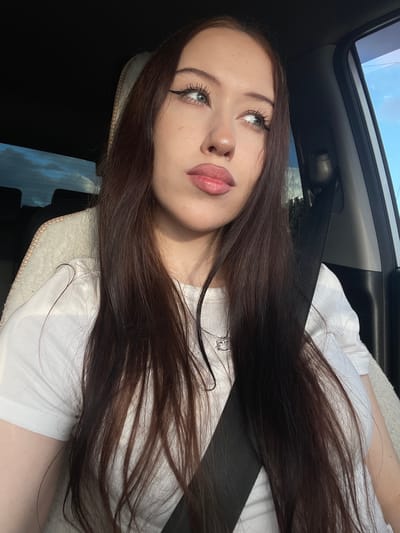 A young woman with long, dark brown hair is captured in a selfie taken inside a car. She is wearing a white t-shirt and a silver necklace with a small charm. Her makeup is prominent, featuring winged eyeliner and lip gloss. She is looking upwards and slightly to the left, with a relaxed expression. The car's sunroof is open, revealing a bright blue sky with some clouds, suggesting it is daytime with fair weather. The interior of the car is visible, including a black seatbelt and a white headrest or cushion. The overall lighting indicates it's likely a sunny day. The image appears to be taken in Gavrilovka, Russia. No text is visible in the media file.