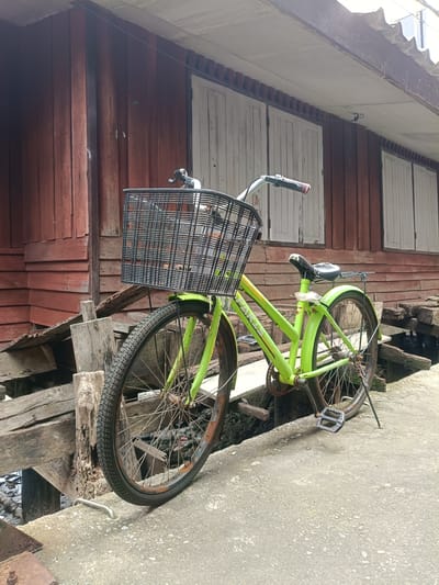 This media file depicts a bright green bicycle parked on a concrete path next to a rustic wooden building. The building has red-stained wooden planks and white shutters on its windows. The bicycle is equipped with a large black wire basket on the front and has a black seat. The wheels and chain of the bicycle show signs of rust, suggesting it may not be new or recently maintained. Wooden planks and beams are stacked and leaning against the building and the path, hinting at construction or disrepair in the immediate vicinity. The ground surface is a rough, grey concrete. The lighting suggests it is daytime, with diffused light indicating an overcast or partly cloudy sky. The scene conveys a sense of rural or semi-rural life, possibly in a tropical or sub-tropical climate given the wooden architecture and the overall atmosphere. The text "PARIS" is visible on the frame of the bicycle. The overall impression is one of a quiet, perhaps slightly weathered, daily scene. The context provided, Tambon Ko Panyi, Thailand, further suggests a coastal or riverside setting, which aligns with the presence of wooden structures that might be elevated or built on stilts.
