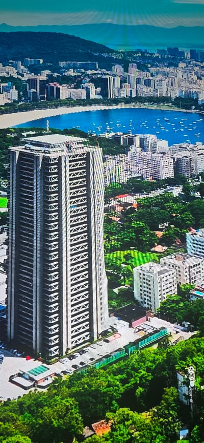 The image displays an aerial view of a city with a large body of water in the background, dotted with numerous boats. In the foreground, a tall, modern skyscraper with a distinctive architectural design featuring a latticed facade dominates the scene. Surrounding the skyscraper are various other buildings, dense green foliage, and a road with parked cars. The overall impression is of a bustling urban environment integrated with natural elements and a waterfront. The image is presented as a digital rendering, exhibiting pixelation and vibrant colors.

The city depicted is not Lippstadt, Germany. The scenery, including the architecture, the presence of a bay with many boats, and the mountainous terrain in the background, is characteristic of a coastal city in a tropical or subtropical region. Lippstadt, Germany, is an inland city with a different geographical and architectural profile.