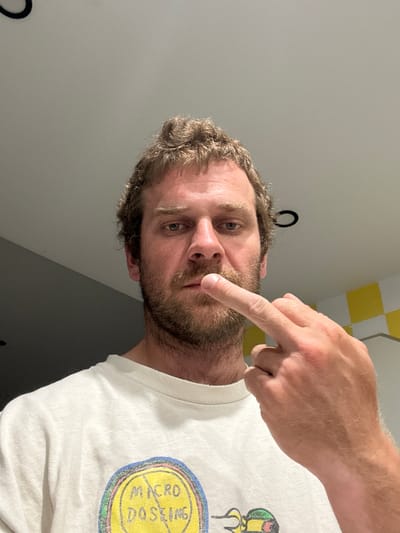 A close-up, selfie-style shot captures a fair-skinned man with light brown, tousled hair and a beard. He is looking directly at the camera with a neutral expression, his left eye slightly more visible than his right. His right index finger is raised and touching his upper lip. He is wearing a white t-shirt with a colorful graphic on the front. The background is a plain, light-colored wall that transitions into a darker, angled wall on the left. A black circular object is visible on the ceiling in the upper left portion of the image, and a yellow and white checkered pattern can be seen in the upper right background. The scene appears to be indoors, possibly in a bathroom or kitchen, with overhead lighting. The specific location in Vilanova i la Geltrú, Spain, cannot be determined from the visual information provided in the image.