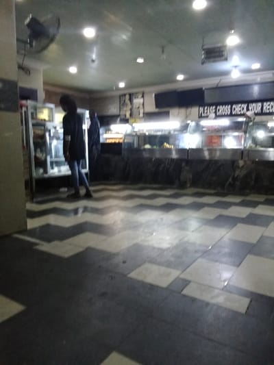 The image shows the interior of a food service establishment in Ovwian, Nigeria, located at coordinates 5.503 latitude and 5.799 longitude.  There are two people in the image. One person is standing near the back of the establishment, facing a glass display case filled with food. The second person is walking toward the display case. The display case has a sign above it that reads "PLEASE CROSS CHECK YOUR RECEIPT."  The floor is tiled with a black and white checkered pattern. There are overhead lights and a ceiling fan in the room. The walls are painted white. The image was taken from the perspective of someone standing near the wall at the left side of the image.