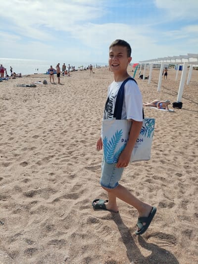 A candid, medium shot captures a young person smiling at the camera while walking on a sandy beach in Blagoveschenskaya, Russia. The individual, a boy with short brown hair, is in the foreground, wearing a white t-shirt with a graphic design, light-wash denim shorts, and dark green slides. He carries a white tote bag with blue leaf designs and text that appears to say "PATRON HOT...RT & SPA .COM.TR". His left eye is slightly squinted, and he has a relaxed smile on his face.

The beach, a mix of sand and small pebbles, stretches behind him. In the mid-ground, numerous people are visible lounging, swimming, and walking along the shore. The ocean glimmers under a partly cloudy sky, with a bright reflection of the sun on the water's surface. To the right, white wooden pergolas line the beach, offering shade. Further in the background, a person in a blue swimsuit is lying on a towel under one of the pergolas. The scene suggests a warm, sunny day at a popular beach resort.