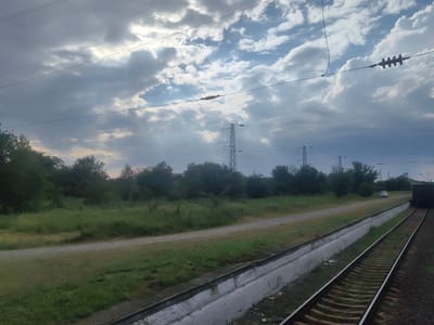 This is an outdoor, daytime, eye-level, medium shot of railroad tracks. The tracks run diagonally from the bottom right to the middle left of the frame. The sky is cloudy with shafts of sunlight breaking through. Several power lines crisscross the sky, supported by tall towers in the background. A dirt road runs parallel to the tracks, separated by a grassy verge and a low concrete barrier. Green trees and bushes line the far side of the road. A white car is visible on the road in the distance, and a dark structure, possibly a building or a freight car, is partially visible at the far right of the frame. The scene appears to be located in an industrial or rural area of Novokubansk, Russia, likely a railroad yard or a line on the outskirts of the city. There are no people visible in the image.