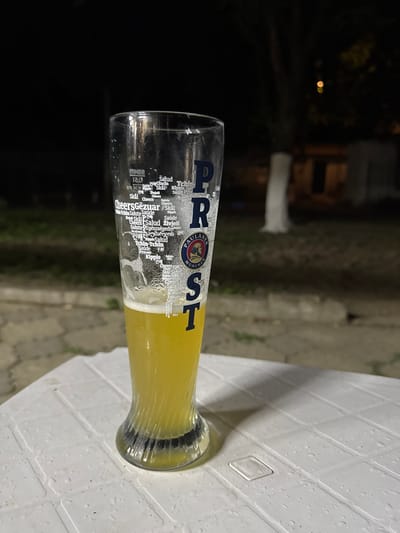 A tall glass of beer sits on a white plastic table outdoors at night. The glass is partially filled with yellow beer and has a frothy head. The glass is decorated with various phrases written in different languages, including "Salud", "Cheers", and "Skål". A logo for Paulaner München is visible on the glass. The background is dark and blurry, with trees and buildings visible in the distance. The table surface is wet, suggesting recent rain. The scene appears to be a casual outdoor setting, possibly a restaurant or bar. There are no people or animals visible in the image. The image was taken in Zugdidi, Georgia.