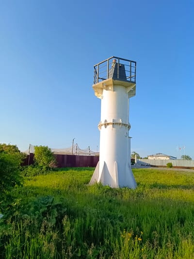 An outdoor, full shot captures a white, cylindrical lighthouse with a black railing around its top, situated in a grassy area under a clear blue sky. The lighthouse is made of metal and has a wider, flared base with a reinforced foundation. A person is visible standing on the platform at the top of the lighthouse, looking out. In the background, a dark red fence runs horizontally, partially obscured by lush green trees and bushes. Beyond the fence, there are some low-lying buildings, flagpoles flying flags, and what appears to be a net structure. The scene is brightly lit by sunlight, indicating it is daytime. The environment suggests a coastal or riverside location in Korsakov, Russia, given the presence of a lighthouse.