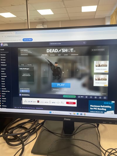 The image presents a forensic view of a computer monitor on a desk within an indoor environment, specified as being in Hérouville-Saint-Clair, France.
The central subject is an iiyama brand computer monitor displaying a web browser interface. The browser is Google Chrome, configured in French, showing the website "crazygames.fr" and specifically the game "DEADSHOT.io." The game's main screen features a stylized male character in tactical gear holding a rifle, positioned above a prominent blue "PLAY" button. Below this, options for "Private" and "Join" are visible, along with "2473 Players Online." To the left of the character, "Daily Challenges" are listed. The webpage also displays several advertisements: a horizontal banner promoting hotel brands (e.g., Radisson, Park Plaza) beneath the game interface, and a partial vertical advertisement on the right side for "DELO Maximum Reliability for PIC Bonding." The Windows operating system taskbar is visible at the bottom of the screen, showing various application icons including a search bar, Microsoft Edge, Google Chrome, File Explorer, and others.
Beneath the monitor, a tangled mass of black electrical and data cables rests on a light-colored desk surface. The background behind the monitor reveals a commercial or institutional setting, characterized by a white suspended ceiling with recessed square fluorescent light panels. A reflection in a surface above the monitor shows a partial view of the room, including what appears to be a white office machine, possibly a printer or copier. No individuals are directly visible in the immediate physical environment of the image, only the character depicted on the screen.
