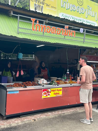 This media file captures a vibrant street food scene in Tambon Wang Prachan, Thailand. The primary subjects are people and the food stall they are interacting with. A man, wearing a light beige t-shirt and light-colored shorts, is leaning over the counter, appearing to order or observe the food. He is dressed casually, suggesting a warm day.
The stall itself is the central object, with a bright green corrugated metal roof and a prominent red facade. A large yellow sign with Thai script and the words "Fried Chicken by Ameena" in English, along with "Pulot Ayam Kahna," indicates the type of food sold. The sign also features the Halal logo, suggesting the food is prepared according to Islamic dietary laws. Below the main signage, a Coca-Cola advertisement is visible, partially obscured.
The scene is busy with activity behind the counter. At least three women, two of whom are wearing hijabs and aprons, are working at the stall. They appear to be preparing or serving food, with trays of fried chicken and skewers prominently displayed. The atmosphere seems bustling and transactional, typical of a popular street food vendor.
Notable details include the various food items, such as skewers of what appears to be fried chicken or meat, displayed in trays. Several bottles of drinks, including Coca-Cola, are visible on shelves behind the counter. The presence of the Halal logo and the mix of Thai and Malay text ("Pulot Ayam Kahna") suggest a diverse cultural context. The ground beneath the stall is paved, indicating an urban or semi-urban setting. The lighting suggests it is daytime, though the exact time of day is not clear. The overall impression is a lively and authentic street food experience in Thailand.