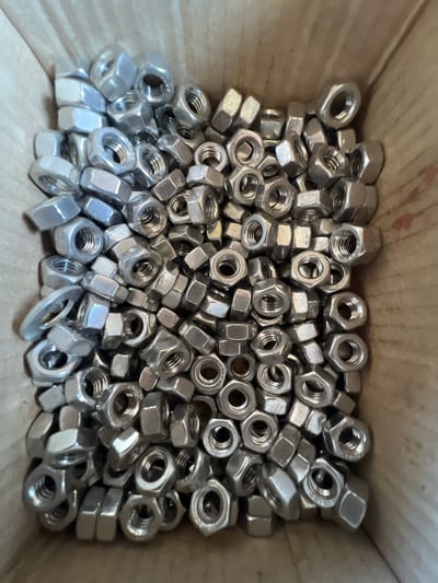 The image shows a pile of hexagonal metal nuts, mostly silver in color, contained within a cardboard box. The nuts are of various sizes and are jumbled together. Some nuts are stacked on top of others, while some are lying on their sides. The texture of the nuts appears smooth and metallic, with visible threading on the inside of some. The box itself is made of light brown cardboard, with some slight wear and tear visible on the edges. There are no people or any specific landmarks indicating a location in Lanžhot, Czechia. The scene is a close-up, overhead shot of the contents of the box.