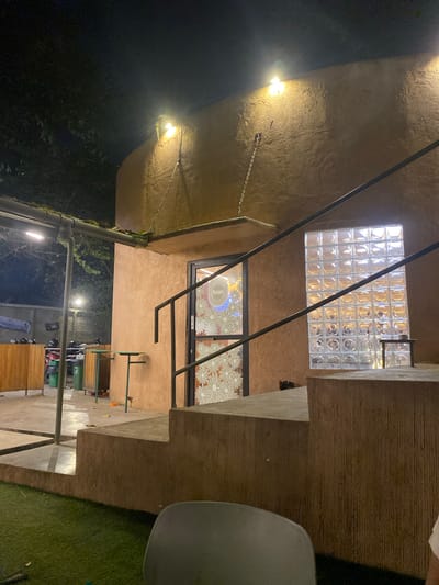 The image captures a dimly lit outdoor scene at night, likely in Bandar Lampung, Indonesia, with artificial lighting illuminating a textured, sandy-colored building facade. A set of concrete stairs leads up to an entrance with a frosted glass door adorned with a floral pattern and a circular logo that reads "bun". To the right of the entrance, a large window constructed of glass blocks emits a warm glow. A black metal railing runs diagonally across the frame, framing the stairs and the building. In the lower right corner of the frame, a dark-colored object, possibly a cat, is resting on a ledge. The surrounding environment includes trees and foliage visible in the background, as well as some tables and what appears to be a seating area. The overall impression is of a quiet evening at a commercial establishment.