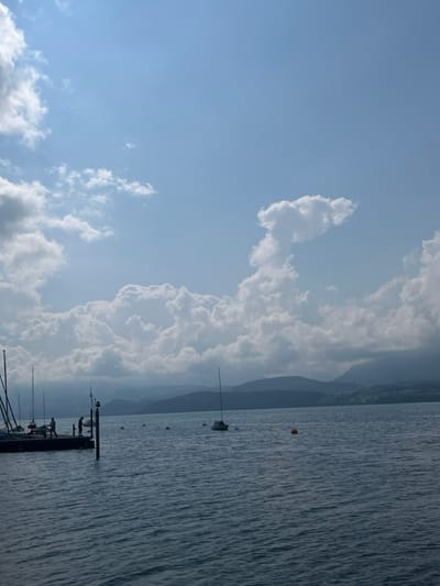 The image depicts a panoramic view of Lake Thun in Hünibach, Switzerland. The sky occupies the upper two-thirds of the frame, featuring a clear blue expanse at the very top that transitions to a broad band of dense, white cumulus clouds across the middle and lower sky. A distinct, vertically oriented cumulus cloud formation, resembling a mushroom cloud or a stylized animal head and neck, is prominent in the upper right quadrant.

The lower third of the image is dominated by the lake's surface, which exhibits gentle ripples and a deep blue-grey coloration. Extending from the left foreground into the water is a dark wooden pier or dock. Three indistinct human figures are present on this pier: one figure is on the far left, appearing to lean or bend; a second figure stands in the middle; and a third figure stands near a dark vertical post at the right end of the pier. Multiple sailboats, identifiable by their visible masts, are moored or docked alongside the pier.

In the mid-ground, a single white-hulled sailboat with a visible mast is independently moored in the open water, positioned to the right of the pier. Several small, white spherical buoys are scattered across the water's surface, and one orange spherical buoy is visible in the mid-right section. The background features a continuous range of dark blue-grey mountains or hills stretching horizontally across the horizon, with patches of green land visible at their base. The scene is illuminated by bright, natural daylight.