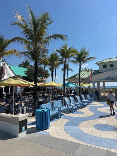 The image depicts a sunny outdoor scene on a pedestrian promenade in Lauderdale-by-the-Sea, United States of America, adjacent to the Atlantic Ocean. The sky is clear blue. Multiple green-fronded palm trees are prominent throughout the frame. The promenade pavement features a distinctive light blue and off-white curvilinear, wave-like pattern.
On the left, a building with white siding and a turquoise-colored roof section houses the "ARUBA BEACH CAFE," as indicated by a partial sign. Its outdoor dining area is furnished with numerous small tables and dark-framed chairs, many with white patterned backs, situated under yellow and light blue patio umbrellas. To the right, a white building with a green, multi-gabled roof and supporting columns displays an American flag mounted on a pole and a sign reading "Relax...you're LAUDERDALE-BY-THE-SEA." A small bird is perched on the roof of this building.
A row of light blue wooden Adirondack-style chairs is arranged on the promenade, facing the visible ocean where several distant vessels are present on the horizon. A blue slatted cylindrical trash receptacle stands on the promenade, near a stone pillar with an attached dark plaque on its left side.
Approximately twelve individuals are visible. Several are seated at the cafe tables under umbrellas, while others stand near the buildings. In the foreground-right, a female subject walks away from the camera, wearing a light-colored long-sleeved shirt and dark shorts, carrying a woven bag over her right shoulder, and holding a leash attached to a small, partially visible dog at her feet.
