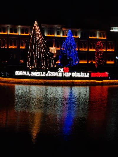 This media file captures a vibrant night scene in Eskisehir, Turkey, showcasing a festive illumination, likely along a river or canal. The foreground features the dark, rippled surface of water, which beautifully reflects the bright lights from above.
In the middle ground, three prominent, conical structures, resembling decorated trees, are covered in string lights. From left to right, these lights are golden-orange, vibrant blue, and red, creating a striking color contrast against the dark surroundings. Below these illuminated structures, a long sign displays glowing text.
In the background, a large building with multiple stories and numerous windows is brightly lit with warm orange and white lights, outlining its facade and windows and adding to the overall luminous atmosphere. The sky above is completely dark, emphasizing the brightness of the artificial lights.
The visible text on the sign reads "2026" above the main phrase "SEVGİYLE, EMEKLE, ÖZENLE HEP BİRLİKLE ESKİŞEHİR". This translates to "WITH LOVE, LABOR, CARE, ALWAYS TOGETHER ESKİŞEHİR," suggesting a message of unity and well wishes, possibly for the upcoming year 2026. The reflections of all these lights are clearly visible in the water, adding depth and a dynamic quality to the image. The scene suggests a public holiday or special event display.