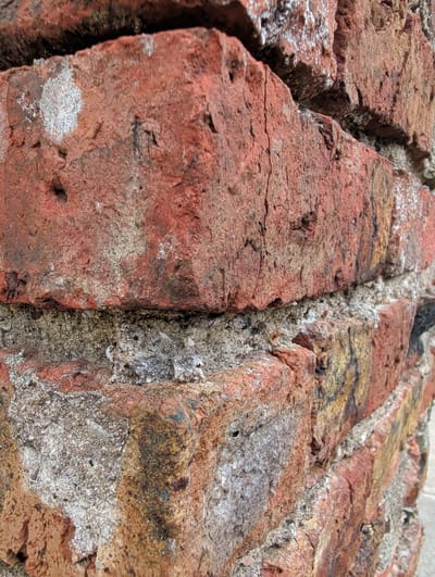 The image shows a close-up view of an aged brick wall, with a focus on the texture and imperfections of the materials. The bricks are predominantly a reddish-brown hue, with variations in color and texture suggesting wear and weathering. Some bricks exhibit patches of white or light grey, possibly mineral deposits or remnants of mortar. The mortar itself appears to be a coarse, grayish mixture, with visible aggregate and signs of erosion or degradation. The overall impression is one of a weathered, possibly historic structure. The location of this specific brick wall within the city of Waterford, Ireland, cannot be determined from the image alone.