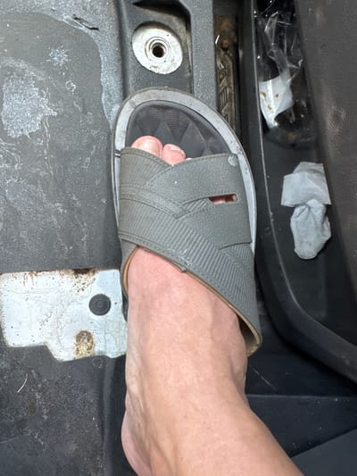 The image shows a close-up view of a person's left foot wearing a gray slide sandal, positioned on the floorboard of a vehicle. The foot and sandal are in the foreground, with the foot partially visible inside the sandal. The surrounding environment appears to be the interior of a car or truck, with a dark-colored textured floor mat and a black plastic trim piece visible to the right. To the left, there is a white metal plate with a circular indent and some rust spots, suggesting it might be part of the vehicle's chassis or a foot pedal. A dirty crumpled white tissue is visible in a crevice of the plastic trim on the right side of the image. The scene is lit by an overhead light source, casting shadows. The location is specified as Knoxville, United States of America.