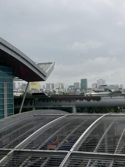 An elevated view captures a section of Hòa Cường Ward, Vietnam, under an overcast sky. In the immediate foreground, the metallic, ribbed, arched roof structure of a building is visible, spanning the lower half of the frame. Below this grid, indistinct forms and equipment are present. To the left, the modern, angular roofline of an adjacent building extends diagonally into the frame, featuring a light grey fascia and a darker underside. Below this roof, a section of a glass-paneled wall is visible.

Mid-ground features an elevated concrete road or pathway, characterized by its curved design and a dense layer of purple-flowering vines cascading from its edge. A dark-colored vehicle, likely a car, is partially visible on this elevated structure, moving from left to right. Support pillars for this structure are also discernable.

The background presents a dense urban skyline composed of numerous buildings of varying heights and architectural styles. Prominent structures include a taller green-glass skyscraper towards the center-right, several white and grey high-rise residential or commercial buildings, and a mix of lower-rise structures. The sky is uniformly grey with diffused cloud cover, indicating an overcast day. No discernible individuals or explicit interactions are clearly visible within the frame.