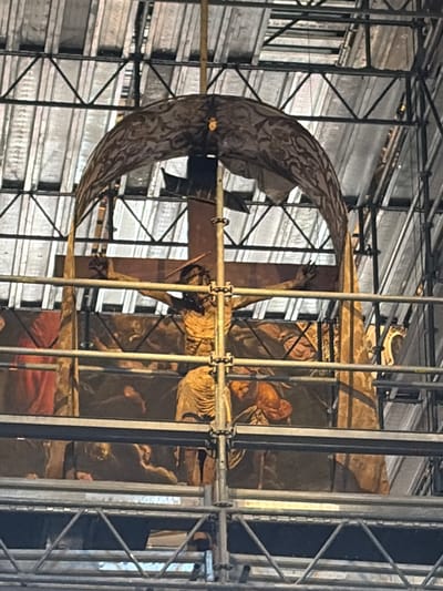 The image shows scaffolding surrounding a religious artwork, specifically a crucifix. The crucifix is positioned in front of a painted panel depicting what appear to be figures in a biblical scene. The surrounding environment is dominated by the metal scaffolding structure.

The location is in Bergamo, Italy.