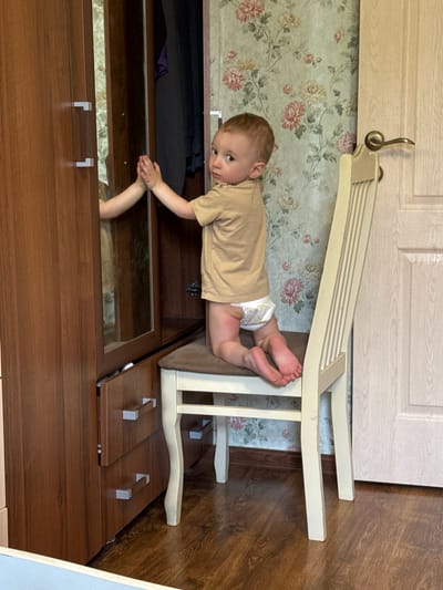 A young child, appearing to be around 1-2 years old, is the central subject of this image. The child is wearing a light brown t-shirt and a diaper. They are lying on their stomach on a piece of furniture, with their feet and legs extended. The child's head is turned to the left, and their gaze is directed towards the camera with a curious expression.

The child is positioned on a light-colored, slatted bench-like object that is propped against a floral-patterned wallpaper. Behind the wallpaper, a light-colored wooden door is visible, with a decorative brass hook on it. The scene appears to be indoors, likely in a home.

Below the bench, a dark brown wooden dresser or cabinet is visible. It has a mirrored surface on its top drawer, reflecting the wallpaper and the child's legs. The dresser has silver handle pulls on its drawers. The floor is covered with a wood-patterned laminate.

The lighting suggests it is daytime, and the overall atmosphere is domestic and casual. The child's pose and expression suggest they are exploring or playing. There is no visible text in the image. The provided location context is Zassovskaya, Russia.