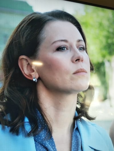 The image is a close-up, slightly angled portrait of a woman with brown hair and blue eyes. She is wearing a light blue collared jacket over a dark blue garment with white polka dots. She has a small, silver earring with a pearl in her left ear. The background is blurred, showing green foliage, suggesting an outdoor setting, possibly in the city of Sloboda, Russia, though this location cannot be confirmed from the visual information alone. The lighting creates a bright highlight across her cheekbone and forehead. Her expression is neutral, and she is looking off to the right.