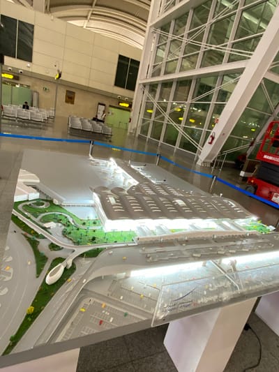 The image shows an airport model, likely representing Houari Boumediene Airport in Dar El Beïda, Algeria. The model is detailed, featuring a terminal building with multiple concourses, a runway area with a miniature airplane, and a parking lot. The surrounding environment of the actual airport is visible in the background, including large windows, seating areas, and what appears to be airport infrastructure like signage and lighting. A red scissor lift is also partially visible to the right, suggesting ongoing construction or maintenance work. The model is placed on a table and is cordoned off with a blue tape barrier. Text on the model base includes "Aeroport d'Alger" and "Algiers International Airport". There are a few people visible in the background, appearing to be waiting or working.