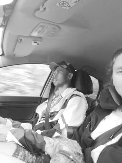The black and white photo depicts the interior of a car, likely in motion as evidenced by the blurred scenery visible through the window. The driver, a male with a beard, wears a cap and a jacket with dark and light stripes. He has his seatbelt on and is looking out the window. On the passenger's lap is a cat wearing a harness. The passenger's face is partially visible on the right side of the frame. The car's interior features include the ceiling light console and sun visor. The location, Малая Уса, Russia, cannot be verified within the context of the image.