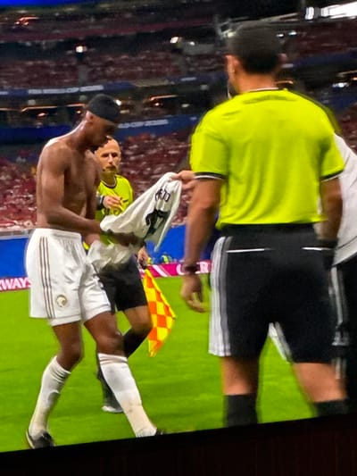 In an outdoor stadium setting, a shirtless Black male soccer player wearing white shorts with a logo on the left thigh and a black beanie is handing a white soccer jersey to a Caucasian male referee. The referee is wearing a bright yellow short-sleeved shirt and black shorts with white stripes. He is holding the jersey which has a number "17" visible on the back. Another Caucasian male, likely a player or official, in a yellow shirt is standing behind the shirtless player. A checkered yellow and red flag is held by an unseen person to the right of the referee, partially visible. The ground is covered in green artificial turf. In the background, a blurred crowd fills stadium seating under stadium lights. The image is cropped at the bottom and sides, obscuring any view of the sky or surrounding structures. No specific details about the city of Bykovo, Russia are present within the image.