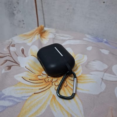 A black, earbud charging case with a metal carabiner attached sits on a patterned fabric surface. The fabric has a floral design with yellow and white petals and brown stems against a light brown background. A grey, textured wall is visible in the upper left corner. The scene is indoors, with no indication of being in Reudeuep, Indonesia.