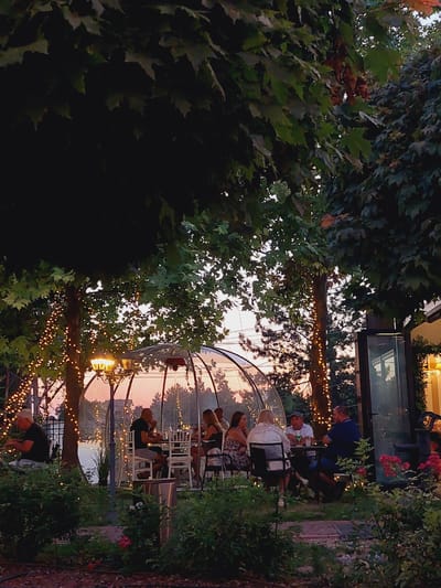 This image depicts an outdoor dining scene at dusk, possibly at a restaurant or a waterfront establishment. In the foreground, lush green bushes and some red flowers are visible, partially obscuring the view. A clear, bubble-like structure, likely a dining igloo or private booth, is set up outdoors. Inside this structure, a group of six people are seated around a table, engaged in conversation and dining. The atmosphere is enhanced by string lights draped around nearby trees, casting a warm glow. The background shows a body of water and a treeline against a soft, dusky sky with hints of pink and orange. The overall impression is one of a serene and intimate outdoor dining experience. The location is identified as Grivitsa, Bulgaria.