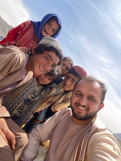 A group of people, seemingly Afghan, is captured in a selfie-style photograph. The foreground features a man with a beard, smiling broadly at the camera. Behind him, several children and an older man are clustered together. The children wear various head coverings, including a blue hijab and a red cap. The older man sports a traditional embroidered hat. All are smiling and appear joyful. The background reveals a bright sky and mountainous terrain, suggestive of a location in a highland area. It is sunny. The location is Ashoqah, Afghanistan. All are dressed in traditional clothing. The photo suggests a spontaneous and cheerful moment captured in an outdoor setting.