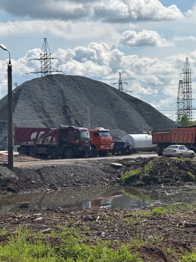 In Kabanikha, Russia, a scene unfolds at what appears to be a construction or industrial site, likely a quarry or material storage area. A massive mound of grey gravel dominates the background, with several towering electricity pylons positioned behind it, hinting at the site's proximity to power infrastructure. In the foreground, two large dump trucks are parked side-by-side: a deep red one on the left, and an orange one to its right. Further to the right, a white trailer and another dump truck are visible. A dark-colored SUV is parked behind the orange dump truck, and a light-colored sedan is parked in front of the rightmost dump truck. The ground in front of the trucks is a mixture of dirt and gravel, transitioning into a muddy, shallow body of water reflecting the sky and surrounding objects. The water is littered with debris, including branches and what appear to be discarded tires. The immediate foreground shows rough ground covered in a dense layer of twigs and low-lying green vegetation. The sky above is partly cloudy with a mix of white cumulus clouds against a light blue backdrop. There are no people visible in the scene. The overall impression is one of an active, if somewhat untidy, operational area.