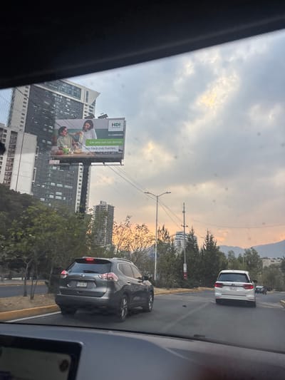 The image depicts an urban street scene captured from inside a vehicle, looking forward through a smudged windshield. The lower portion of the frame shows the vehicle's dark dashboard and the top edge of an infotainment screen.
In the midground, a multi-lane asphalt road is visible. A dark gray Nissan X-Trail SUV with license plate 779-ALY is positioned directly ahead in the left-center lane, facing away from the viewer. Further ahead in the right lane, a white Honda Odyssey minivan (license plate NWN-32-0) also proceeds away from the viewer. Other vehicles are faintly visible further down the road. The road is bordered by a yellow-painted curb on the left and dense green foliage on both sides.
In the background, a tall, modern multi-story building with a dark facade and glass windows dominates the left side of the skyline. A large billboard is centrally located in front of this building. The billboard displays an advertisement for "HDI Seguros" with the text "TU COMPAÑÍA Ser uno con los demás, nos hace más fuertes," featuring two women appearing to share a meal. Numerous other buildings, some appearing to be under construction with visible framework, are interspersed among trees and utility poles with overhead wiring. Mountains are discernible on the distant horizon to the right. The sky is partially cloudy, exhibiting patches of yellow and orange light, indicative of either sunrise or sunset.
The scene is located in Santiago Yancuitlalpan, Mexico.