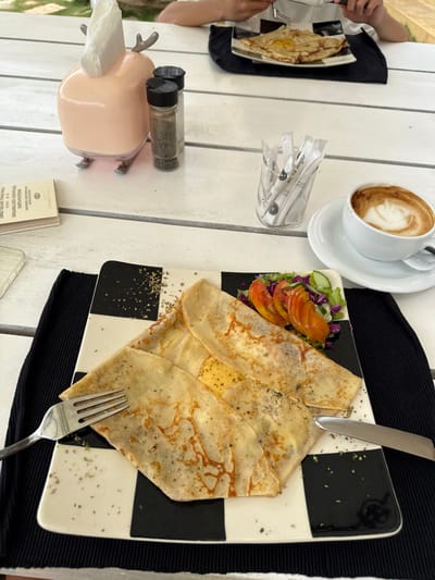 A table setting displays crepes on a black-and-white checkered plate with a side salad. Silverware rests on the plate and nearby. A cup of coffee sits on a saucer, with packets of sugar in a clear container nearby. Pepper and salt shakers are positioned on a pink napkin holder. A second plate with a crepe is visible in the background.