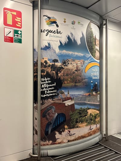 The image depicts the interior of a public transport vehicle, likely a train car, located in Sant Quirze del Vallès, Spain. A large, vertically oriented advertisement poster for "Noguera Turisme" dominates the right side of the frame, spanning from near the ceiling to the floor. The poster is a collage of scenic landscapes and activities, featuring snow-capped mountains, rural villages, a lake or reservoir, and lush green hills.
Within the poster's imagery:
* The top section includes a logo with a stylized bird or creature and the text "Noguera Turisme" with the website "www.turismenoguera.cat". Several smaller institutional logos, including "CONSELL COMARCAL DE LA NOGUERA" and "FGC", are present at the top right.
* A circular inset image on the upper right displays a stone building, possibly a castle or historic structure, surrounded by cypress trees.
* Activities depicted include a yellow paraglider with two figures below it, and three individuals on bicycles riding along a dirt road in a lower section.
* Key promotional terms in black text on a textured background include "Natura," "Aventura," "Allotjament," "Gastronomia," "Patrimoni," and "Experiencies" (Nature, Adventure, Accommodation, Gastronomy, Heritage, Experiences).
* A prominent illustration of an older man with a smiling expression, wearing a flat cap and a striped shirt, is situated in the lower-left quadrant of the poster.
To the left of the poster, a white interior wall displays three safety pictograms: a red square with a yellow fire extinguisher symbol (Escala de socors), a green square with a white downward staircase symbol, and a green square with a white first aid symbol (Camilla de socorro). A silver-colored metal vertical pole, likely a handrail, runs along the left edge of the poster, extending from top to bottom. The floor shows a dark, ribbed grating design on the right, transitioning to a light gray, textured surface on the left. No individuals or direct interactions are visible outside the poster.