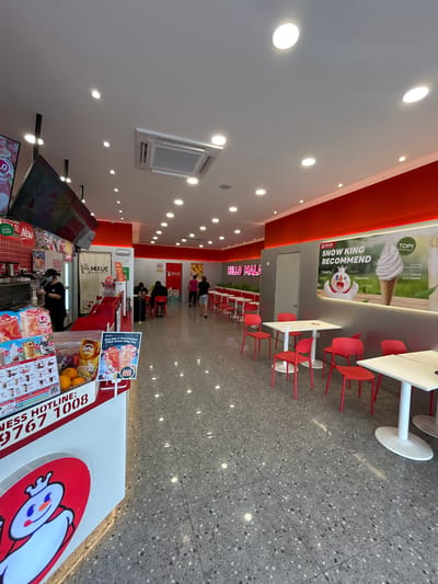 This image captures the brightly lit interior of a Mixue ice cream and beverage shop, likely located within the Subang Jaya City Council area in Malaysia. The shop features a modern, clean design with white walls, a red accent strip along the ceiling, and numerous recessed spotlights illuminating the space. The floor is a grey terrazzo-style tile with speckled patterns.
In the foreground and midground, a prominent service counter is visible. It's adorned with the Mixue snowman character logo and displays various menu items, including "Milk Tea," "Peach Bobo," and fresh fruits like oranges. An employee wearing a black uniform and mask is seen working behind the counter. Digital screens on the counter also display menus and promotions, with text like "UPGRADED!" and a "Grab Food" logo.
To the right, a seating area features several white tables with red bases and matching red chairs, arranged for customers. Further back, customers can be seen seated or waiting.
Prominent wall signage includes a large poster advertising "MIXUE ICE CREAM" as a "TOP1 MIXUE Product" with "400,000,000 Sold," and featuring the "SNOW KING RECOMMEND" character described as "{tasty} {playful} {flavorful}". Another sign on the wall reads "HELLO MALA". Additional text visible includes promotions like "Buy Any 2 New Product FREE Snow King Toy" and a "BUSINESS HOTLINE: 9767 1008". An air conditioning unit is mounted on a side wall. The atmosphere appears casual and inviting, typical of a fast-food or dessert establishment. The ample lighting suggests it is daytime.