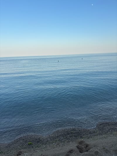 This is an eye-level, high-angle photograph of a beach and the sea, oriented vertically. The scene depicts a tranquil coastal landscape at what appears to be either dawn or dusk, judging by the soft, diffused light and the presence of the moon in the clear blue sky on the left side of the frame.

The left portion of the image is dominated by a vast expanse of pale blue sky, gradually transitioning to a lighter, almost white hue near the horizon. A faint crescent moon is visible in the upper left quadrant of the sky.

The middle section of the image shows the calm, blue sea, characterized by gentle ripples and subtle variations in color, reflecting the sky. Two dark, indistinct objects, possibly buoys or small boats, are floating on the water, positioned vertically in line with each other.

The rightmost part of the frame features a dark, pebbly beach. The pebbles are small and densely packed, with some sparse green vegetation visible amongst them. Footprints are clearly impressed into the sand and pebbles, suggesting recent human activity. The water gently laps at the shore.

There is no visible text in the image. The overall mood is serene and peaceful, evoking a sense of quiet solitude. The location is identified as Osenovo, Bulgaria.