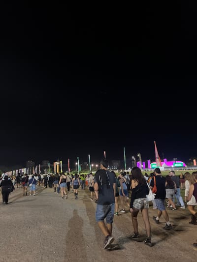 The scene depicts a large outdoor gathering at night, specifically at what appears to be the Hipódromo de San Isidro in Martínez, Argentina. The ground consists of a wide, unpaved dirt path, illuminated by an unseen light source that projects elongated shadows of people onto the ground. The sky overhead is dark and devoid of visible stars.
A dense crowd, comprising an estimated 50-70 individuals, moves away from the camera and towards the background. The majority of participants appear to be young adults dressed in casual attire, including shorts, t-shirts, and light tops.
In the immediate foreground, a male subject is visible from behind, wearing a dark t-shirt with a light-colored garment draped over his left shoulder, blue denim shorts, and sneakers. To his right and slightly forward, a female subject is also viewed from behind, wearing a black off-shoulder top, camouflage-patterned shorts, and fishnet stockings with dark footwear.
The background features multiple tall, vertical flags or banners in colors such as green, white, pink, and orange, lining a distant area. Beyond these, several large, illuminated structures emit bright pink/magenta and green light, suggesting stages or display screens. White fences, characteristic of a racecourse, are visible on the far right side of the background. Distant architectural structures and trees are faintly discernible against the dark horizon to the left.