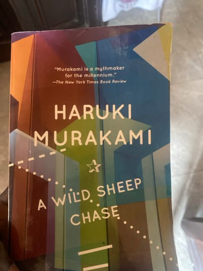 An indoor, close-up shot captures the paperback cover of the book "A WILD SHEEP CHASE" by Haruki Murakami. The book, slightly angled, rests on a light-colored, soft surface, possibly fabric. The cover features an abstract design composed of geometric shapes in shades of blue, green, brown, yellow, and white.

Visible text on the cover includes the author's name, "HARUKI MURAKAMI," and the title, "A WILD SHEEP CHASE." A quote, ""Murakami is a mythmaker for the millennium." -The New York Times Book Review," is also prominently displayed. The top left corner of the book shows minor wear. No people or specific activities are depicted. The lighting suggests an indoor setting, but the time of day and weather are not discernible from the image itself. The provided context indicates the location is Lakka, Sierra Leone, though no visual cues in the image confirm this.