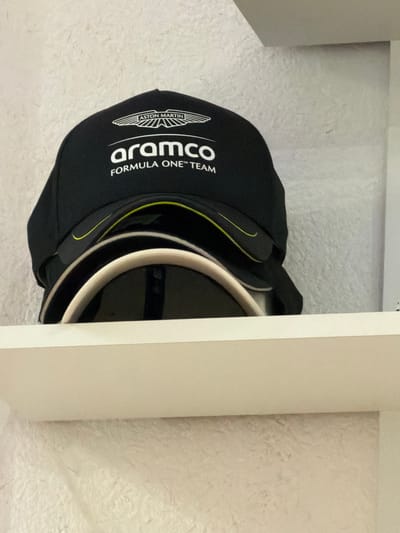 A close-up, angled shot shows a black baseball cap with the "Aston Martin Aramco Formula One Team" logo resting on a textured white surface, partially obscured by a vertical, light-colored surface that appears to be part of a drawer or cabinet. The cap features a white brim with a contrasting black band and a thin yellow accent line. The image is taken indoors, but the specific location in Kovel, Ukraine, cannot be determined from the available visual information.