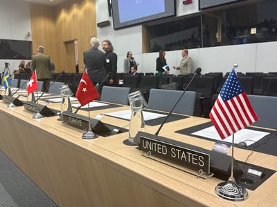 The image shows a conference table with flags and nameplates for Sweden, Switzerland, Turkey and the United States of America. The table is made of light-colored wood and has a black microphone on each side. The chairs in the background are black. Several people are in the background, but their faces are not visible. The coordinates (50.879, 4.422) place the scene in the European Parliament building in Brussels, Belgium.
