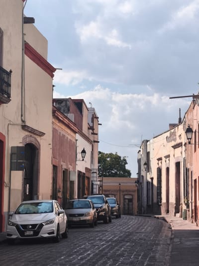 The image captures a street scene in the city of Querétaro, Mexico. A narrow street, paved with cobblestones, curves slightly to the right, lined with buildings on either side. These buildings, characteristic of Querétaro's architecture, feature a variety of pastel shades, with red trim around windows and roofs. Several cars are parked on the left side of the street: a white Nissan Versa, a silver sedan, and a dark-colored SUV. The buildings display ornate details, including wrought-iron balconies and traditional street lamps. The sky above is partly cloudy.