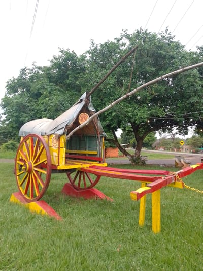 This outdoor photograph features a brightly painted, traditional-style ox cart positioned on a well-maintained grassy area. The cart is the central subject, predominantly colored in vivid yellow and red. It has large, spoked wooden wheels and a wooden bed, topped with a rustic roof made from a combination of corrugated metal and thatch or straw. Two long poles extend forward from the front of the cart, and a metal chain is visible near the front support structure.

The cart itself is elevated slightly on supports that are also painted in yellow and red, blending with its overall aesthetic. In the background, lush green trees with dense canopies create a natural backdrop. Beyond the trees on the right side of the image, a paved road or street is visible, along with some distant buildings and a yellow diamond-shaped road sign, which appears to indicate a speed bump or uneven road ahead. Overhead, utility lines traverse the light, overcast sky.

The scene is set during the daytime, under what appears to be a cloudy or overcast sky, resulting in soft, diffused light without harsh shadows. There are no people visible, and the cart appears to be a static display rather than actively in use. The vibrant colors of the cart suggest a decorative, celebratory, or cultural artifact. The location is Concepción, Paraguay.