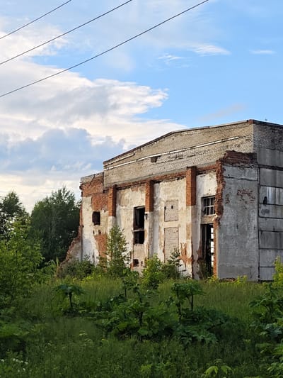 The image shows a dilapidated brick and concrete building, likely an industrial structure or factory, in a state of disrepair. The building's facade is crumbling, revealing exposed brickwork and concrete. Several windows are broken or boarded up, and the roofline appears damaged. The building is surrounded by overgrown vegetation, including tall grass and trees, suggesting an abandoned or neglected location. Power lines are visible in the upper left portion of the frame. The sky is partly cloudy with blue patches. The scene is set in an outdoor environment during daylight hours. The provided information states the location is in the city of Суда, Russia.