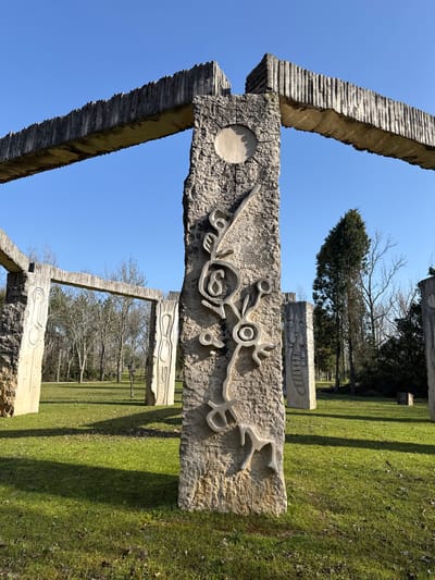 The image depicts the "Monumento ao Foral" (Charter Monument) within the Caneiro Leisure Park in Mealhada, Portugal, under a clear blue sky. A large, textured, light grey stone pillar dominates the right foreground, featuring intricate bas-relief carvings. These carvings include a circular sun motif at the top, radiating lines, and a complex abstract design of interwoven organic and geometric shapes, one resembling the numeral "6". This pillar supports a heavy, horizontally positioned concrete beam with a rough, corrugated texture, extending across the upper portion of the frame and connecting to other structures.
In the mid-ground and background, additional similar stone pillars and horizontal beams are visible, forming an open-air sculptural complex. Some pillars appear connected, creating archway-like elements, while others stand individually. Many of these background structures also feature less distinct abstract carvings.
The ground is covered with green grass, exhibiting variations in color and density, with prominent long shadows cast by the monuments, indicating bright sunlight from the left. Deciduous trees, mostly bare of leaves, populate the mid-ground and background, suggesting a winter or early spring season. A tall, slender evergreen tree stands to the right of the central pillar. A dense line of trees forms the distant background horizon. No human figures or active elements are present in the scene.