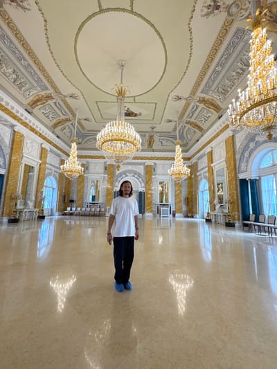 In Strelna, Russia, a man stands in the middle of a grand ballroom with a high, ornate ceiling and two large crystal chandeliers. The room features marble floors that reflect the light from the chandeliers and windows. The walls are decorated with columns, arches, and intricate artwork. The man is wearing a white t-shirt, dark pants, and blue shoe covers. He is holding a phone in his right hand and looking at the camera with a slight smile. The ballroom appears to be part of a palace or historical building.