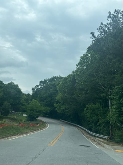 The image depicts a curving asphalt road in Mountain Park, United States of America. The road has double yellow lines indicating the center. A metal guardrail runs along the side of the road. Trees densely border the road. The sky is overcast with clouds.