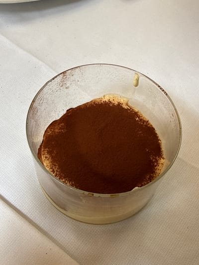 The image shows a close-up of a serving of tiramisu in a clear glass dish, placed on a white tablecloth. The tiramisu is topped with a generous dusting of cocoa powder. The glass dish has some condensation on the exterior. The background consists of the textured white tablecloth, with a diagonal shadow visible to the left of the dish. There is no information in the image to indicate the location in the city of San Giovanni, Italy.