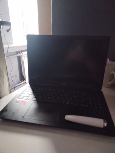 The image shows a black laptop computer placed on a white table. The laptop is open, and its screen is dark. A white cylindrical object with a red tip is lying on the keyboard. To the left of the laptop, a white radiator and a window are visible. A white mug with a handle is on the right side of the table, behind the laptop. The scene appears to be indoors, likely in a room with natural light coming from the window. There is no indication of the specific location within the city of Chita, Russia, as the image lacks any identifiable landmarks or contextual cues.