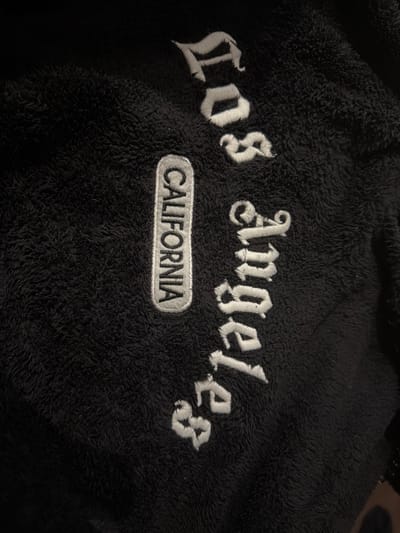 The image depicts a black, furry garment with the embroidered words "Los Angeles" in white, ornate lettering. Below this, a gray patch with black lettering reads "CALIFORNIA." The garment's texture is fuzzy and plush.