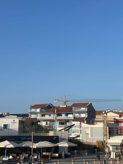 In Gafanha da Nazaré, Portugal, a construction crane is prominently visible against a clear blue sky, its yellow arm extending horizontally over a cluster of buildings. The buildings feature red-tiled roofs and a mix of blue and white facades. Below the buildings, an outdoor seating area is set up with white umbrellas providing shade over tables and chairs. A sign for "bella barra" with "PIZZERIA - GELATINA - LOUNGE BAR" is visible, suggesting a restaurant or cafe. Several people are present in the seating area and walking along a boardwalk in the foreground. The scene depicts a coastal town with ongoing construction.
