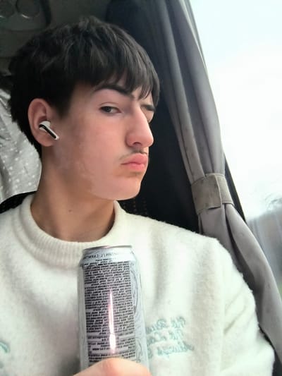 The image captures a young male individual within an interior setting, likely a vehicle, in Mlada Gvardia, Bulgaria. The subject is positioned facing the camera, with his head slightly tilted right, displaying a neutral expression with pursed lips and a faint mustache. He has short, dark hair and prominent dark eyebrows. A white wireless earbud is visible in his left ear. His left cheek displays lighter skin patches. He is wearing an off-white, textured knit sweater with light blue embroidered text visible on the lower right side. The subject's right hand holds a tall, silver-colored beverage can with printed text, including the term "L-CARNITINE," vertically oriented on its surface. To the right of the subject, a grey fabric curtain is drawn and secured by a strap, with a blurred, bright, overcast sky visible through an adjacent window. A light-colored, patterned fabric is partially visible on the left side of the frame, behind the individual's head.